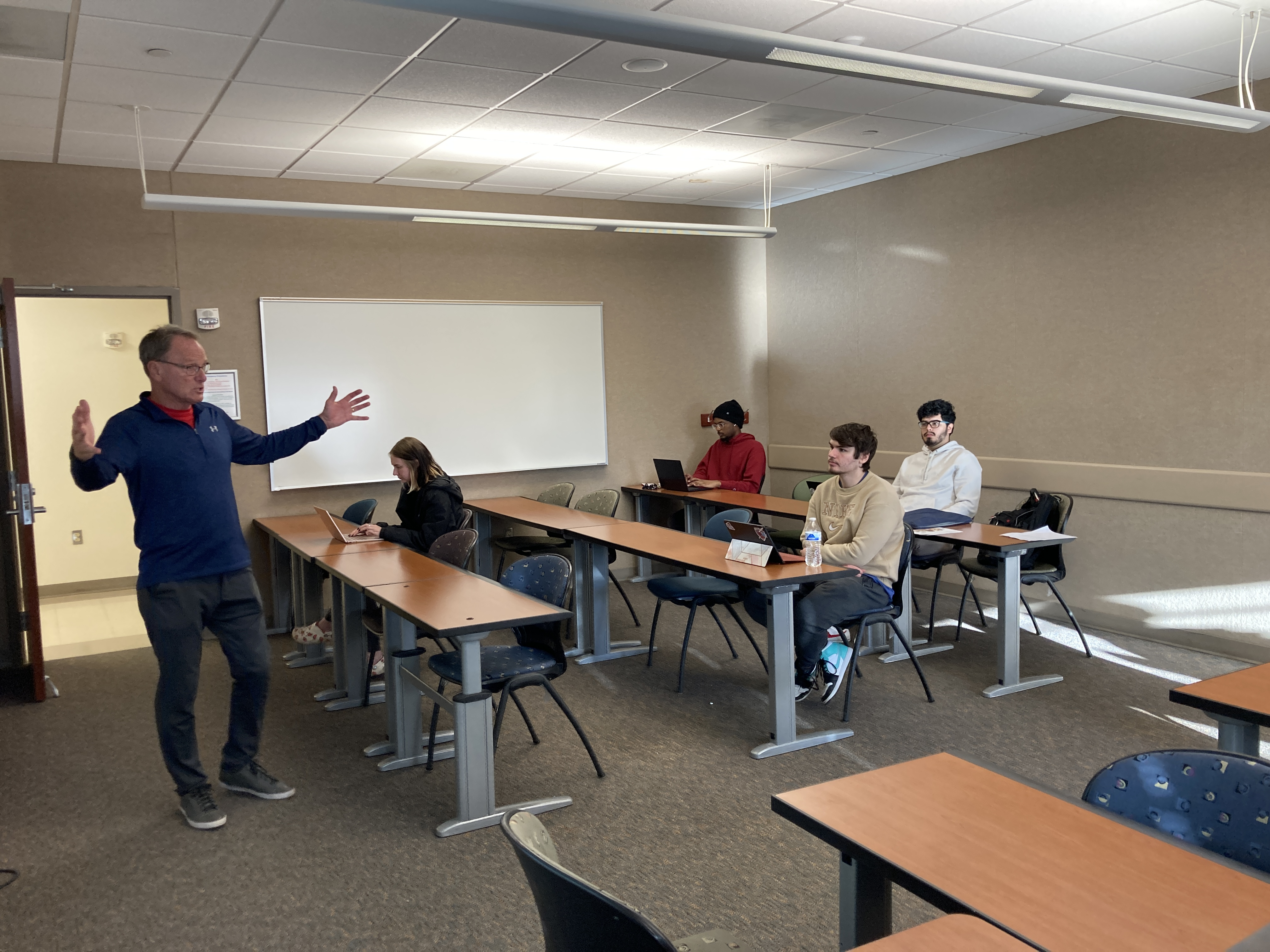 Students in a classroom setting, listening to a speaker with his arms stretched out. 