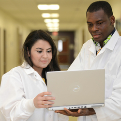 two healthcare informatics students in lab coats looking at a laptop in a hallway