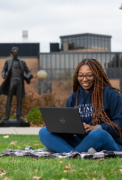 A student sitting on the grass on the UIS quad with a laptop computer with a statue of Abraham Lincoln and the HSB Building in the background.