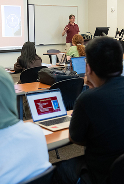 The backs of heads of students sitting in a classroom listing to a professor teach. Several laptop computers are visible.