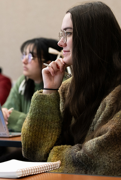 A student in class smiling with a notebook on the desk and pen in hand.