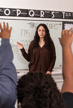 A teacher is shown in a classroom in front of a marker board. Several children's hands are raised to ask a question. 