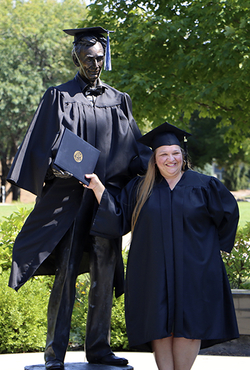 A student in cap and gown stands next to the Abraham Lincoln statue on campus (also in cap and gown).