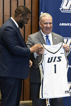 Mike Hermann holding a basketball jersey reading "UIS 1" stands next to Vice Chancellor Jamarco Clark on stage.