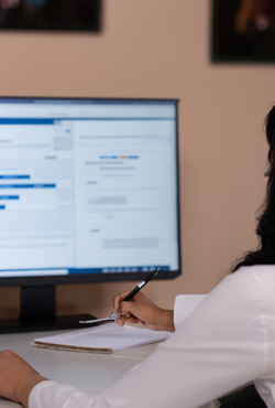 A person from behind sitting at a desk and holding a pen in front of a blurred computer screen.