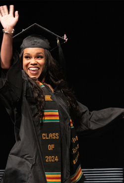 A female UIS graduate waves to the crowd in her cap and gown while walking on stage to collect her degree.