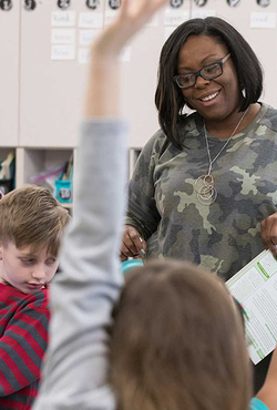 A teacher in a classroom smiling while students raise their hands.