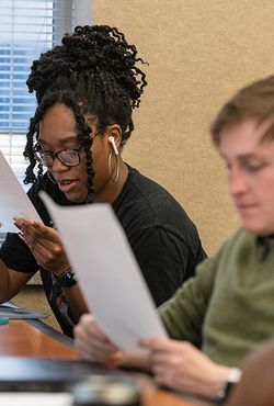 Two students sitting at a desk in a classroom reading pieces of paper. 