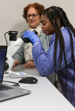 Student using a microscope in a lab while a professor observes, with cell images displayed on a nearby laptop screen.