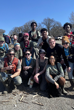 A group of volunteers poses for a photo during an outdoor cleanup event, wearing work gloves, boots, and bandanas. A damaged boat and debris are visible in the background.