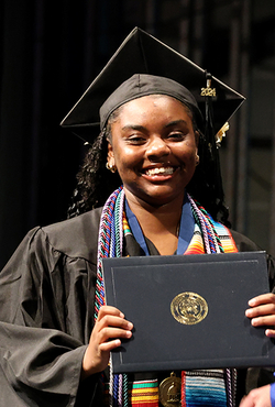 A UIS graduate smiles while holding her diploma on stage during the commencement ceremony, wearing a cap and gown.