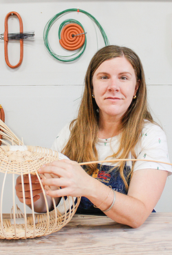 A woman with long hair works on weaving a wicker basket at a wooden table. Behind her, colorful, abstract wall art creates a creative atmosphere.