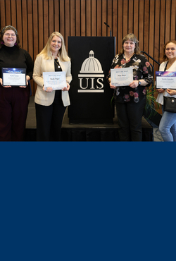 Four award recipients pose with their certificates in front of a UIS podium at a recognition event.