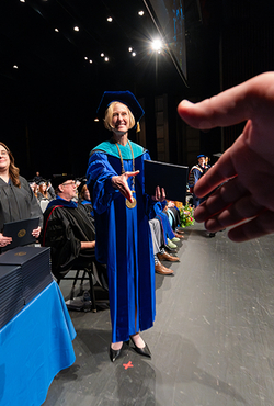 A university official in academic regalia extends a diploma and handshake to a graduate during a commencement ceremony.
