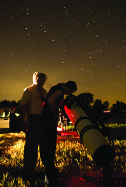Two people using a telescope to stargaze in a field at night under a star-filled sky.