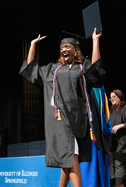 Graduating student celebrates on stage in cap and gown holding diploma at UIS Commencement as faculty applaud.