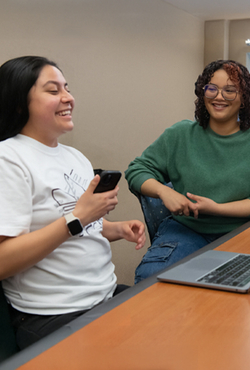 Students laugh and talk in a classroom setting with laptops on desks and a relaxed, collaborative atmosphere.