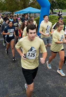 A group of runners participates in a race, starting under a blue inflatable arch. Participants wear numbered bibs and light-colored shirts.