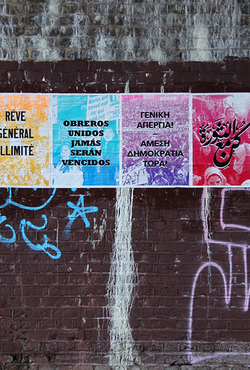 A vibrant poster series on a graffiti-covered brick wall. Each colorful panel displays multilingual text.