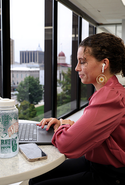 Young woman works on a laptop at a high-rise office window with earbuds, water bottle, and phone nearby.