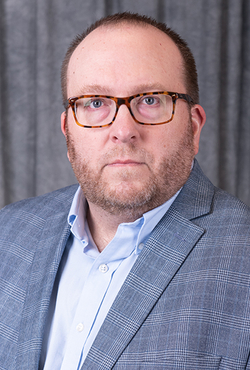 Man in glasses wearing a blue blazer and shirt, against a dark curtain backdrop.