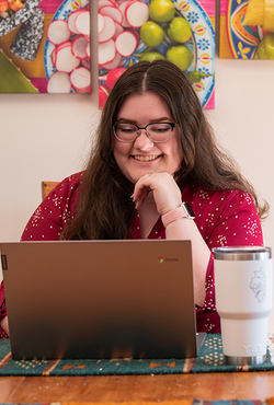 Woman smiling at a laptop with a large mug on the table; colorful art in the background.