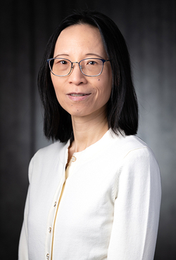 Woman with glasses and a white cardigan, dark background, neutral expression.