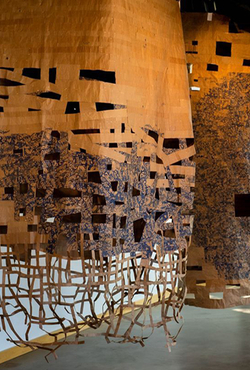 Hanging woven art installation with cutouts, warm tones, near windows.