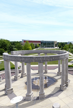 A circular stone monument with tall columns, inscribed with "University of Illinois at Springfield," set in a sunny, green campus.