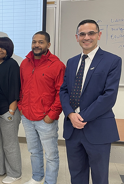 Group of people standing in a classroom, one in a suit, others in casual attire.