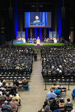 Graduation ceremony with speaker on stage, audience in caps and gowns.