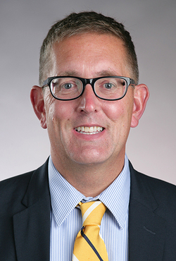 Man in suit with glasses and a yellow tie, smiling against a gray background.