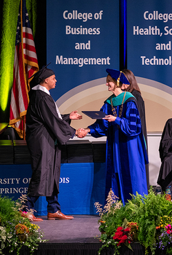 Graduate receives diploma on stage from faculty member in a blue robe.
