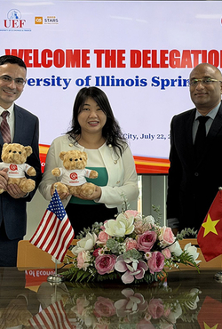Three people smiling, holding teddy bears, with flags and flowers on a table.