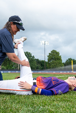 Baseball coach helps player stretch leg on field, cloudy sky.