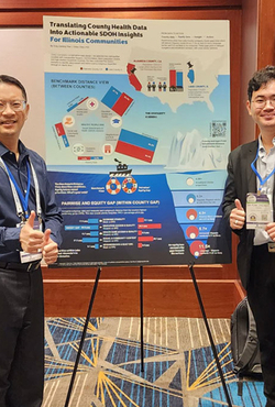 Two men smiling and giving thumbs up, standing beside a colorful conference poster on a stand.