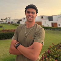 Young man smiling with arms crossed, standing in a garden at sunset.