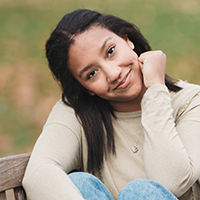 Smiling woman sitting outdoors in casual clothing, hand on chin.