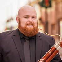 Christopher Raymond in a suit holding a bassoon, smiling outdoors.