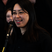 Smiling woman speaking at a microphone, wearing glasses.