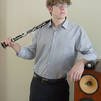 Young man in glasses holding a clarinet, standing next to a speaker.