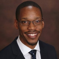Smiling person in a suit and glasses against dark background.