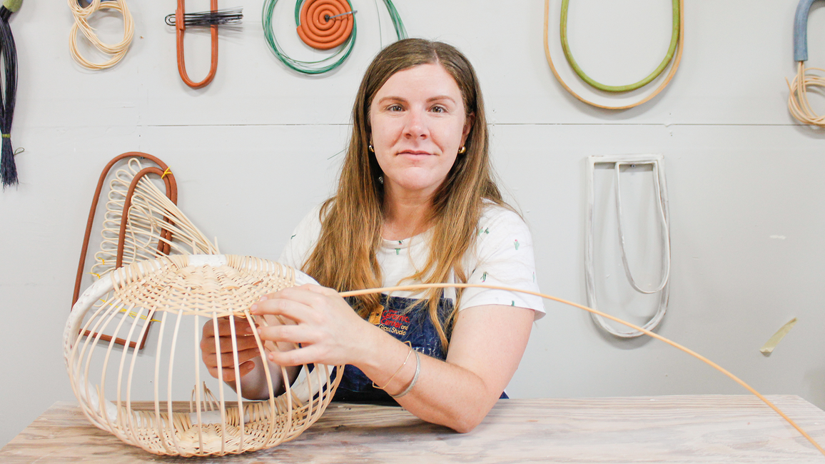 A woman with long hair works on weaving a wicker basket at a wooden table. Behind her, colorful, abstract wall art creates a creative atmosphere.