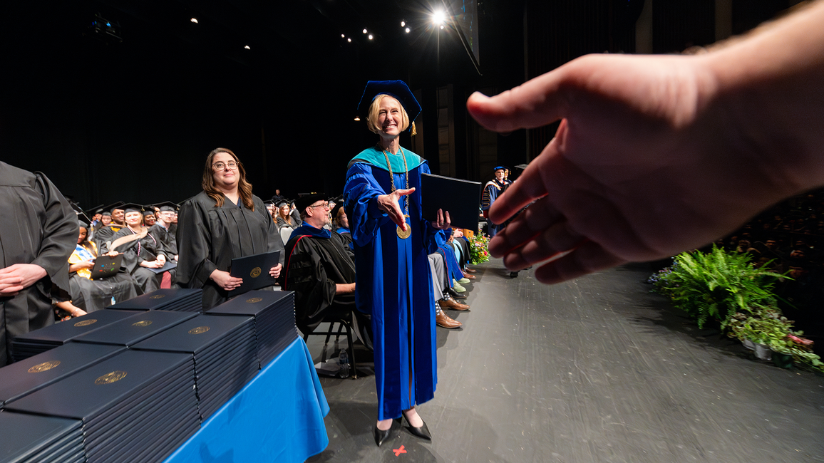 A university official in academic regalia extends a diploma and handshake to a graduate during a commencement ceremony.