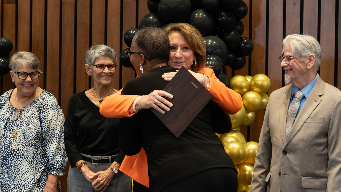 A person receives an award and a hug during the Good as Gold Volunteer Awards ceremony, with others smiling nearby.