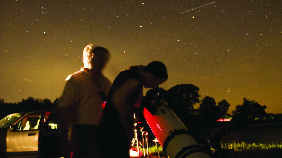 Two people using a telescope to stargaze in a field at night under a star-filled sky.