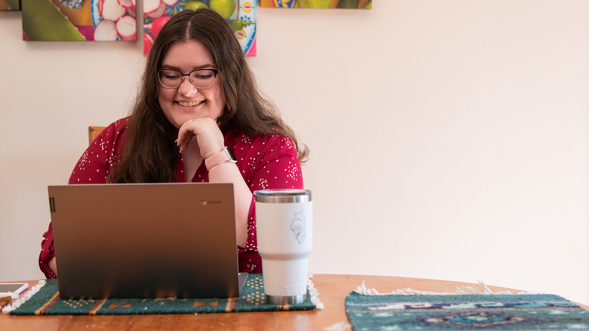 Student smiling while working on a laptop at a table with a tumbler, colorful art hanging on the wall behind her.