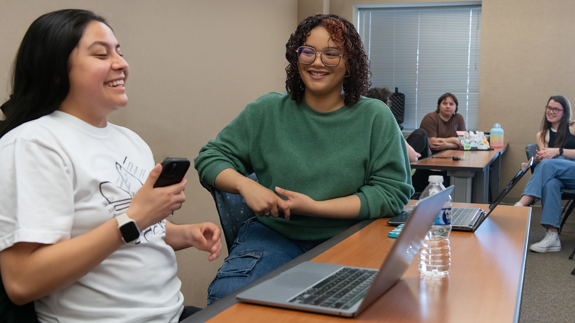 Students laugh and talk in a classroom setting with laptops on desks and a relaxed, collaborative atmosphere.