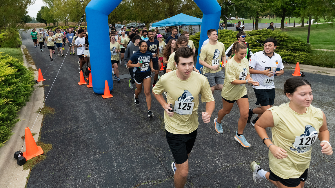 A group of runners participates in a race, starting under a blue inflatable arch. Participants wear numbered bibs and light-colored shirts. The atmosphere is energetic and lively, set against a backdrop of trees and grass.