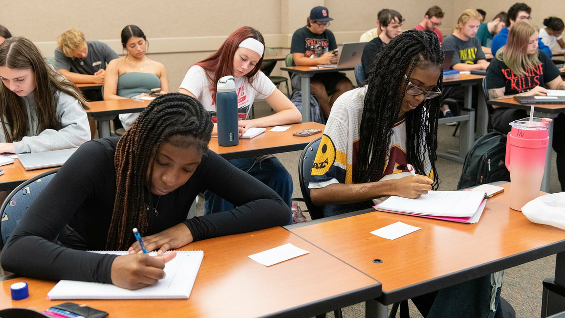 A classroom of students writing in notebooks, laptops open, and water bottles on desks.
